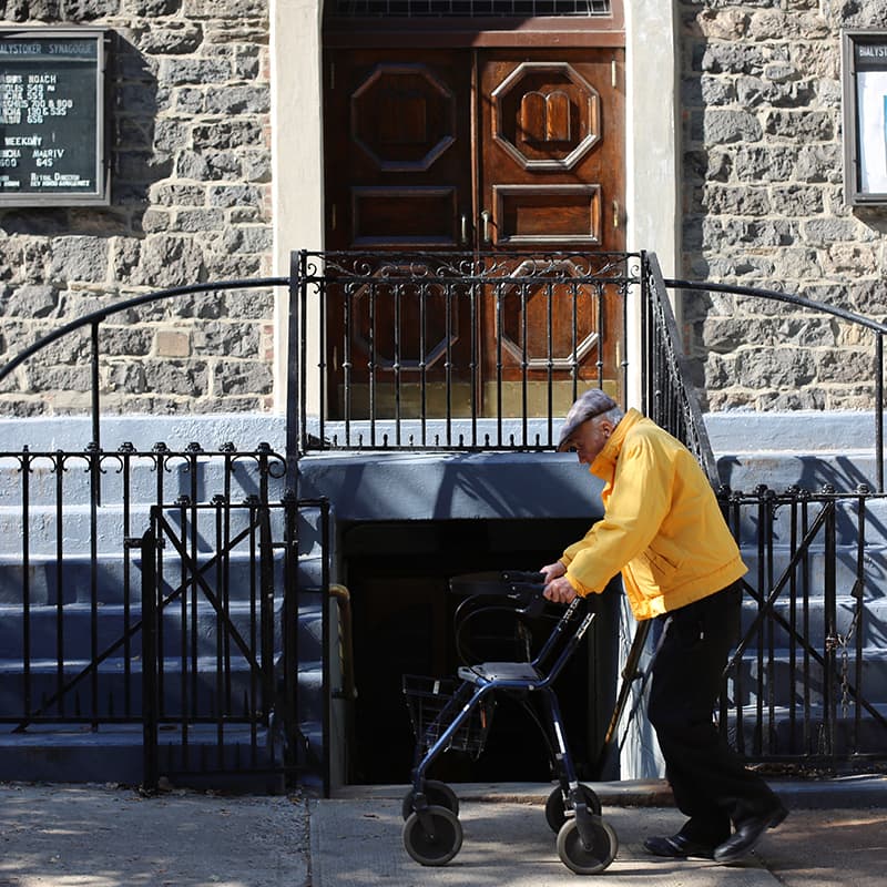 An older man in a yellow jacket using a walker to move past an old building
